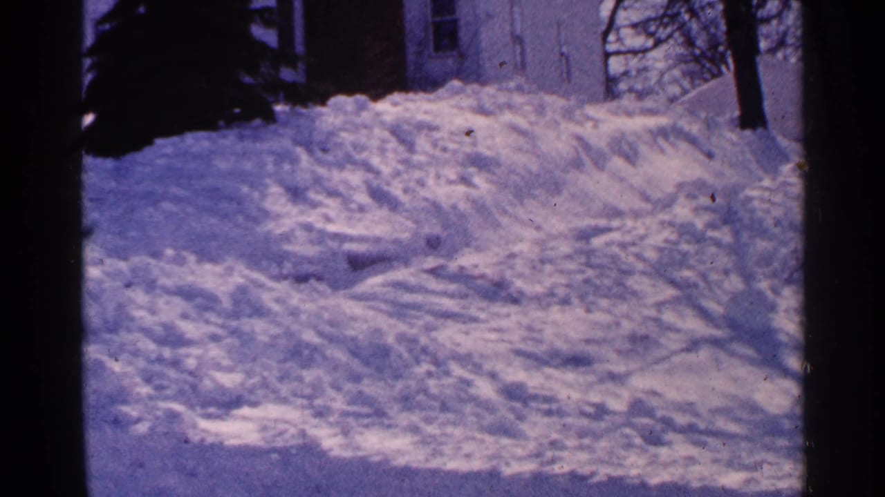 Snow-covered driveway edge with high plowed banks, trees, and a partial house behind.