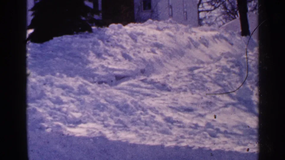 1965 Minnesota 8mm frame with piled snow dominating the yard or driveway edge, backed by a partial residence and winter trees.