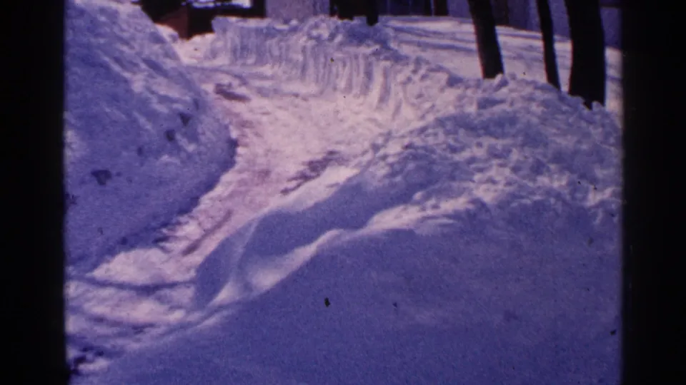 1965 Minnesota home-movie frame showing a narrow cleared snowy path or driveway between tall snowbanks, with bare trees and a residential background.