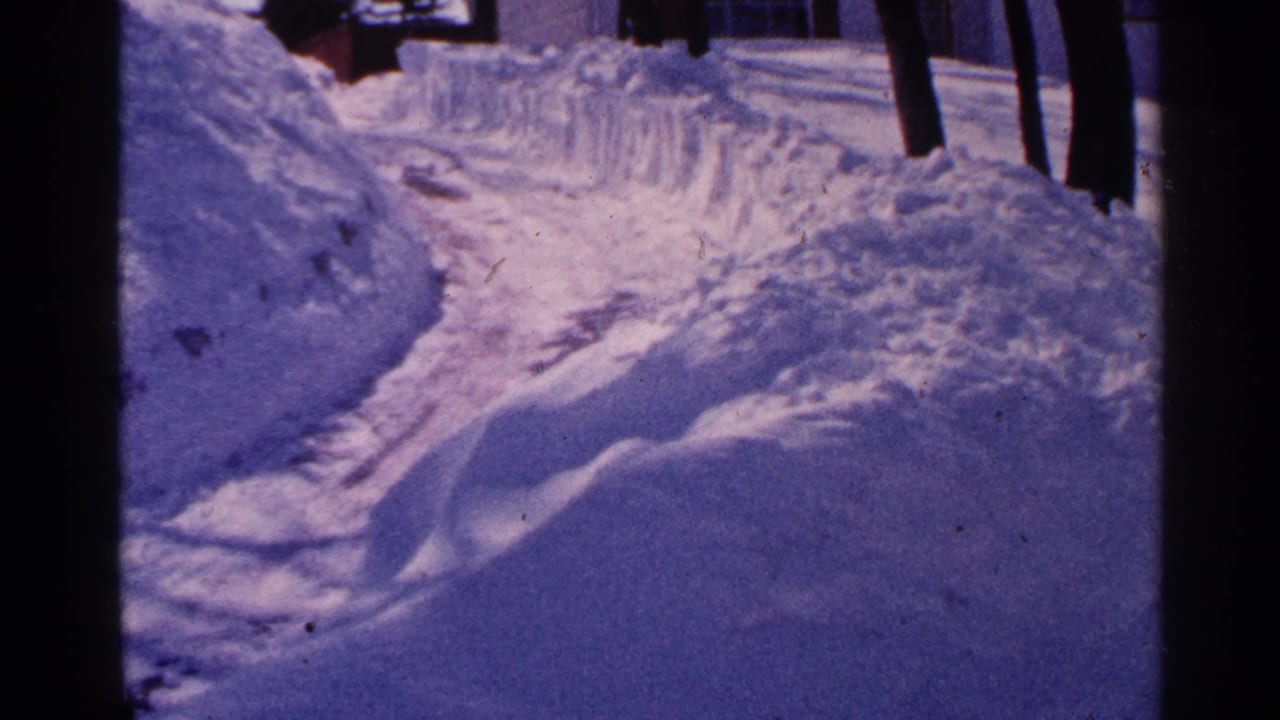 1965 Minnesota home-movie frame showing a plowed snowy residential path bordered by deep snowbanks and bare tree trunks.