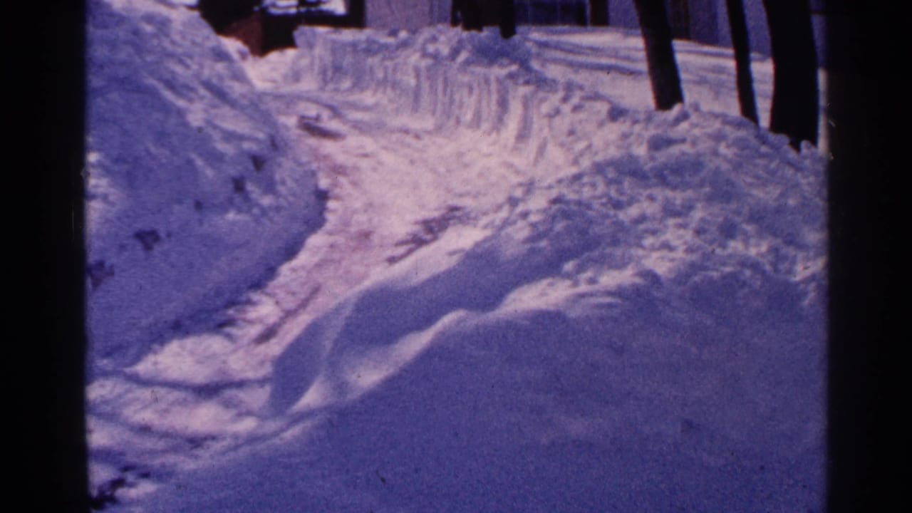 A 1965 Minnesota winter view shows a curving plowed path between tall snowbanks, with trees and a building edge beyond.