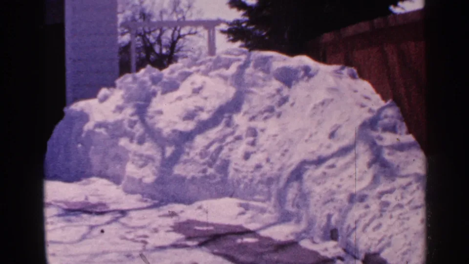 A 1965 Minnesota home-movie frame shows a large roadside snowbank beside a cleared surface, with a fence and house edges nearby.