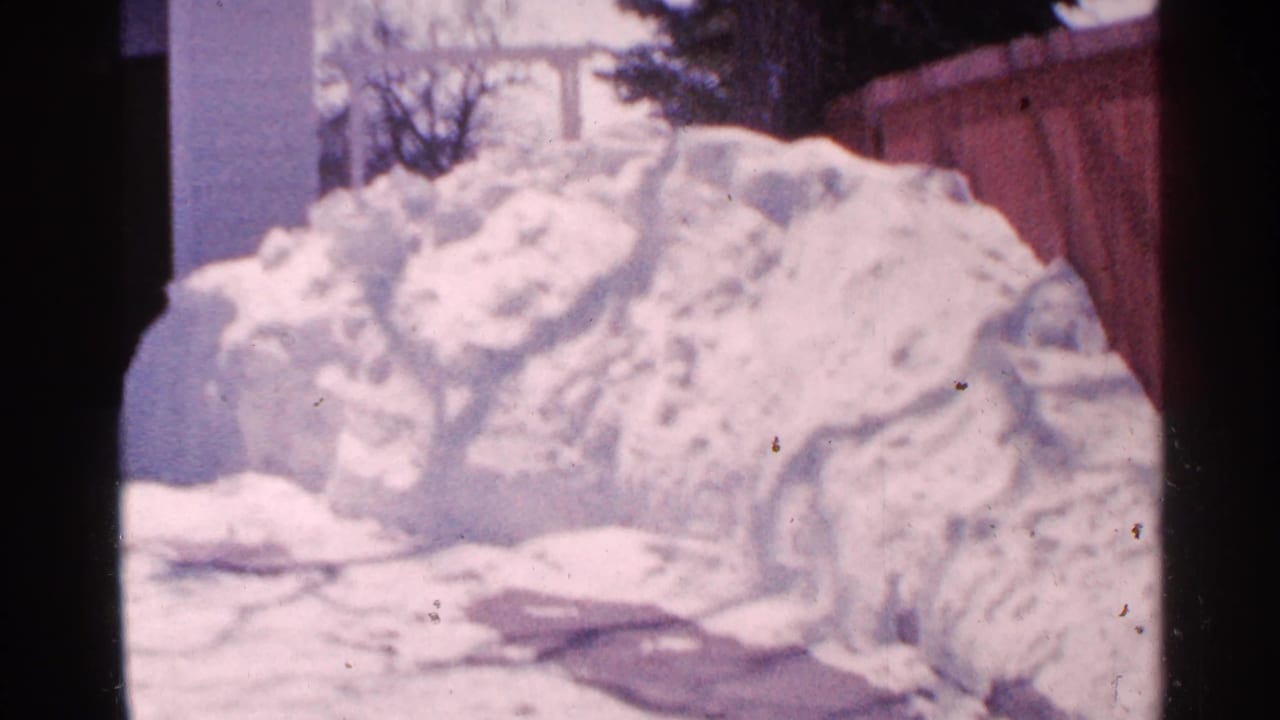 Minnesota 1965 home-movie frame showing a large snowbank in a snowy yard with a wooden fence and house exterior in the background.