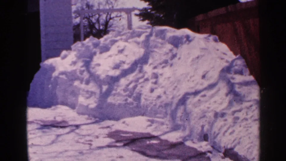 A 1965 Minnesota winter yard view shows a high snowbank, wooden fence, and house exterior with a cool purple-blue film cast.