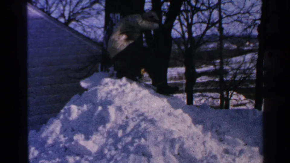 A distant person in a dark coat and hat shovels snow beside a house in metadata-supported 1965 Minnesota winter footage.