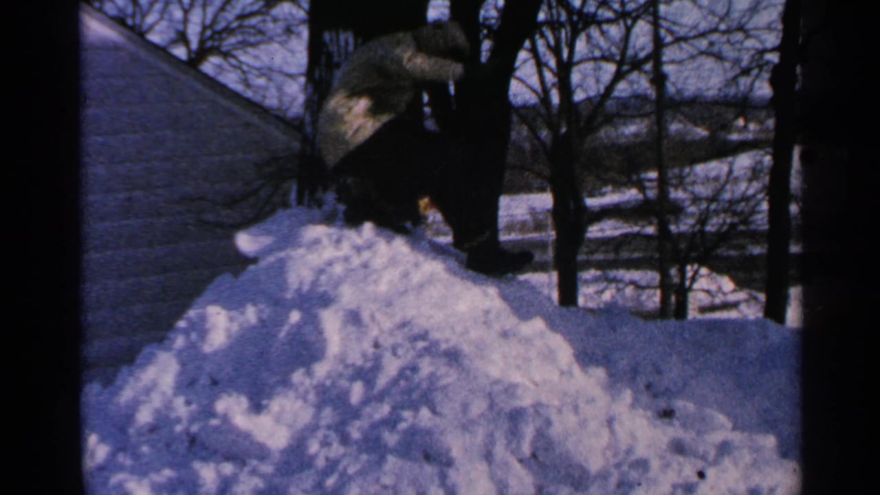 Bundled person crouches atop a large snow pile near bare trees and a house roof.