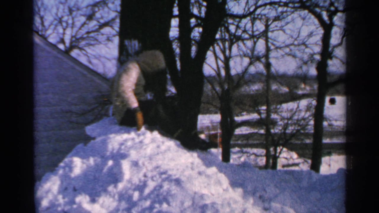 Person in winter clothing bends over a snow pile with trees and a roof behind.