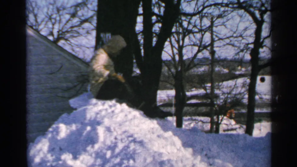 1965 Minnesota 8mm still of a person on a snow mound, arm lowered toward the snow, with a wintry yard and roofline visible.