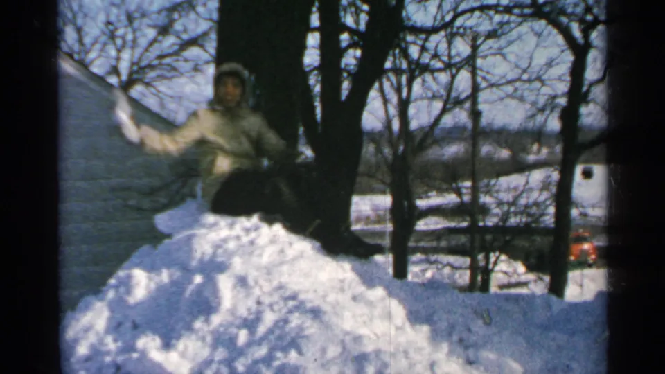 1965 Minnesota winter home-movie frame with a bundled person seated on a snow pile, arm lifted toward the camera.
