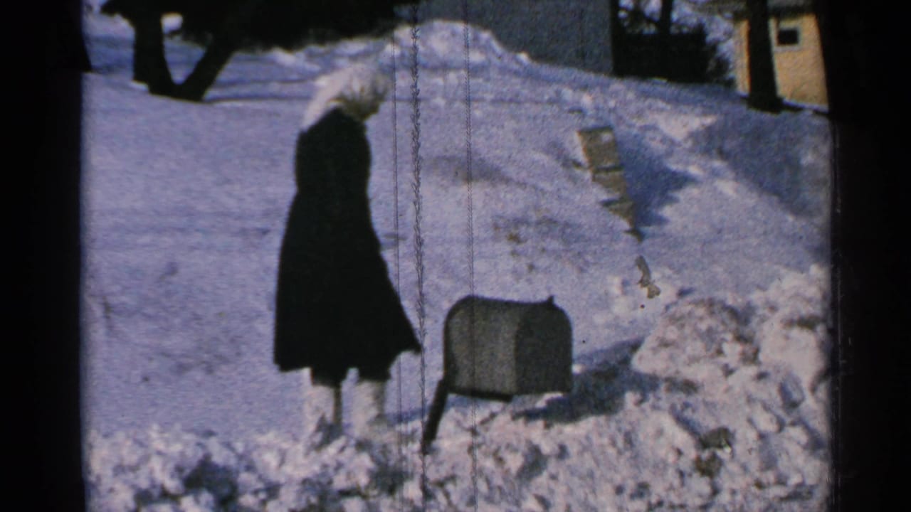 In 1965 Minnesota home-movie footage, a woman in a dark coat approaches a partially buried mailbox in a snow-covered yard.