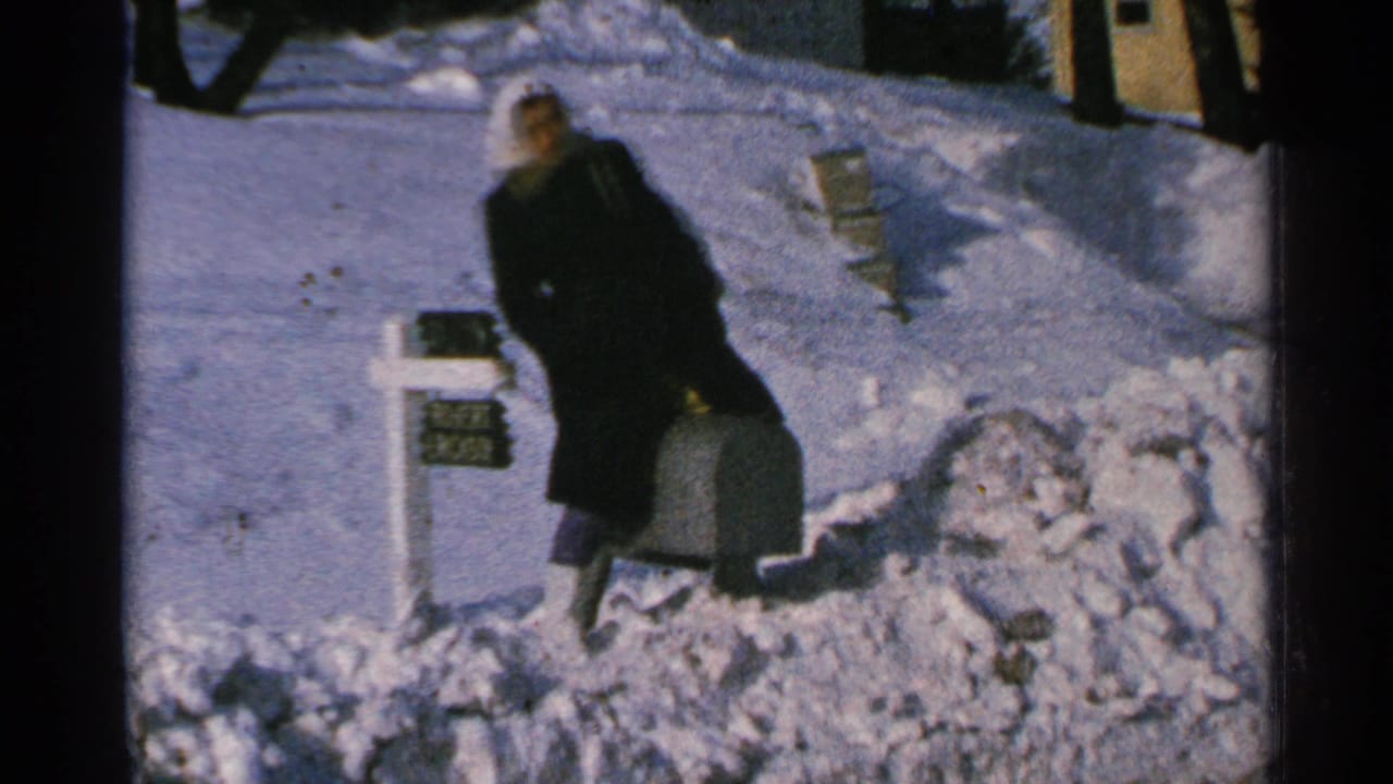 A woman in winter clothing stands next to a mailbox and suitcase amid deep Minnesota snow in 1965 8mm footage.