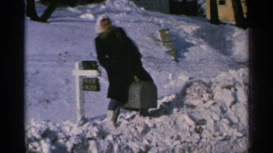 A woman in winter clothing stands next to a mailbox and suitcase amid deep Minnesota snow in 1965 8mm footage.