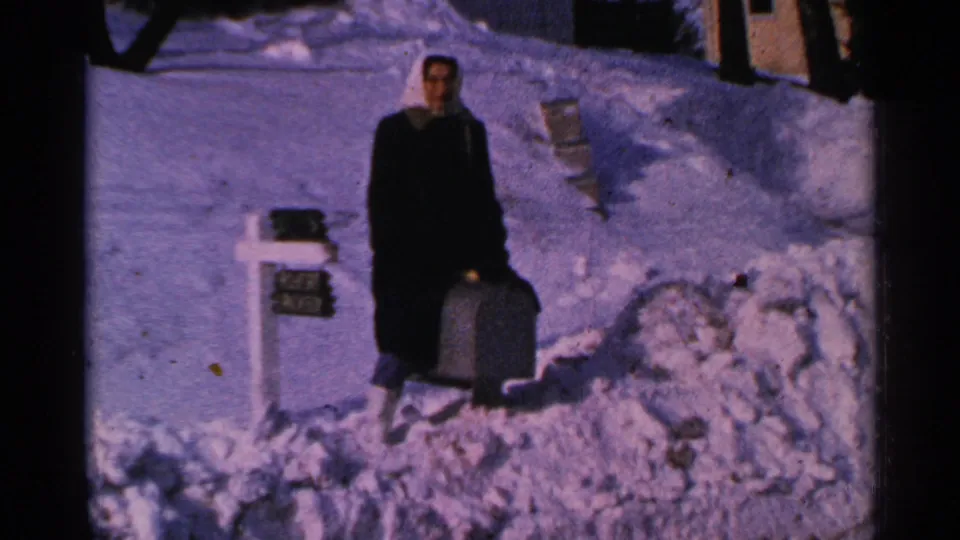 The woman appears to sit or crouch beside the snow-covered mailbox, with a suitcase nearby in the 1965 Minnesota yard scene.