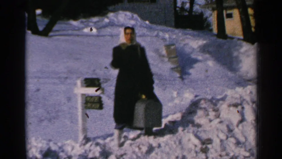 A woman in a dark coat faces the camera beside a buried mailbox and suitcase in 1965 Minnesota winter home-movie footage.