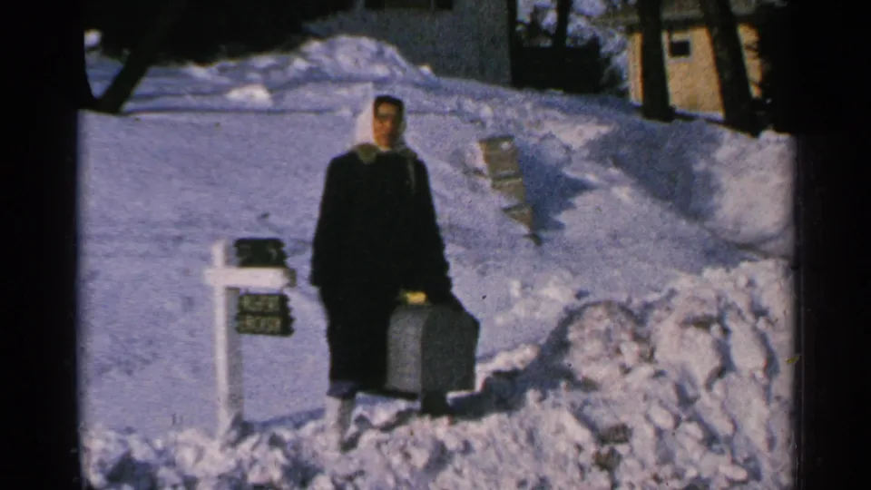 In 1965 Minnesota home-movie footage, a woman stands by a white mailbox amid snowbanks while holding a dark suitcase.
