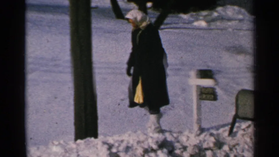 The woman walks away from the snowy mailbox area in 1965 Minnesota footage, with a dark pole crossing the foreground.