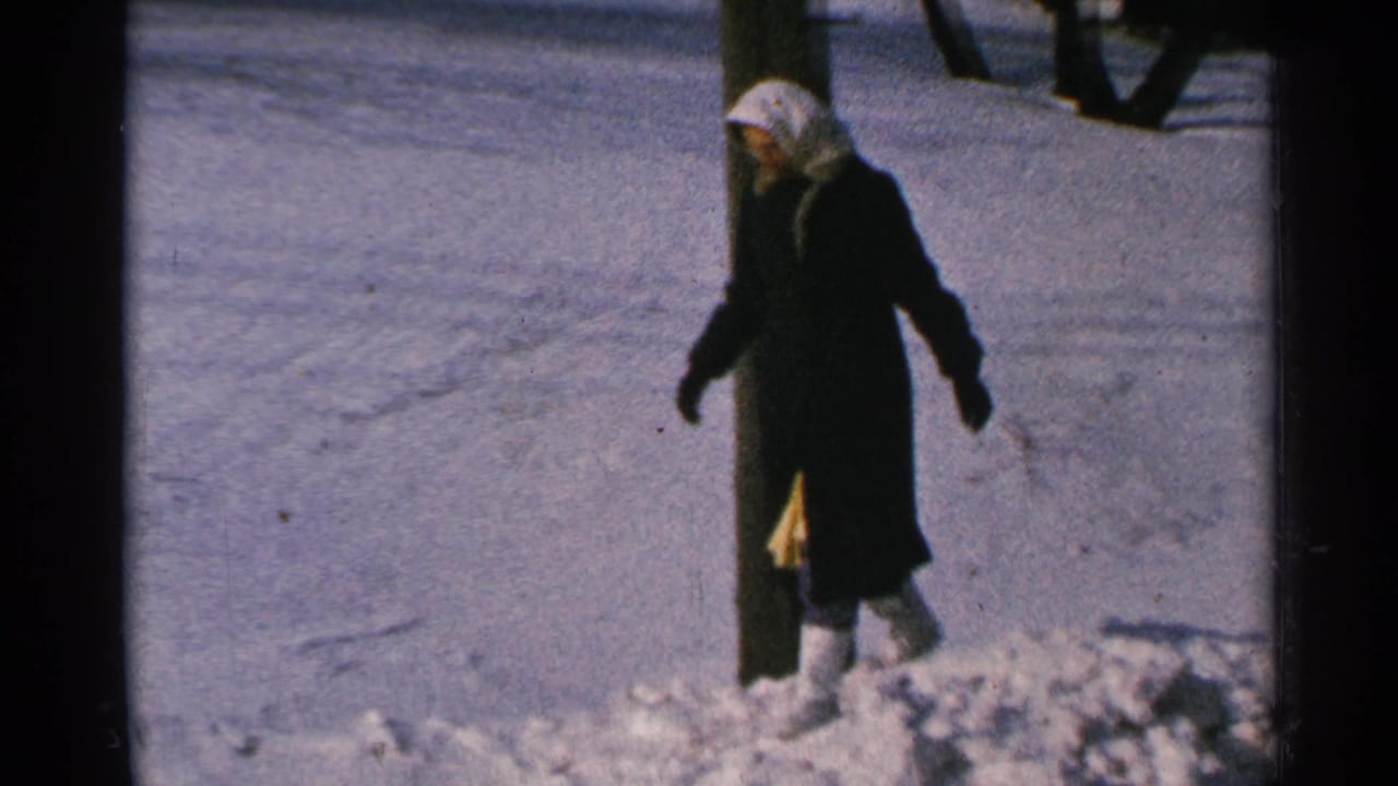 Woman in a dark coat steps through deep snow near a wooden post.