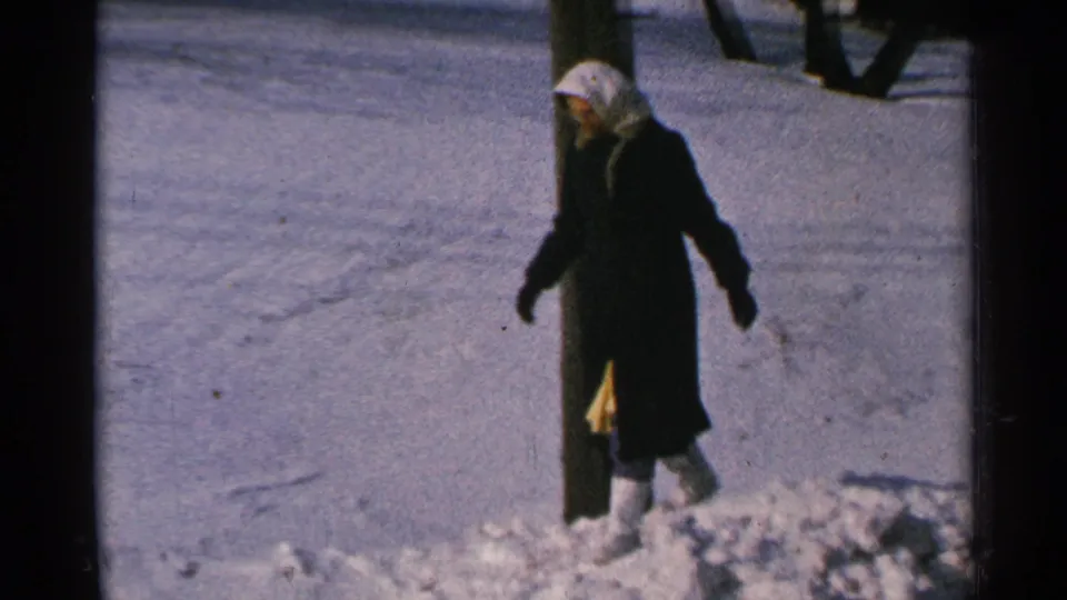 In 1965 Minnesota home-movie footage, a woman in a dark coat and light head covering walks through deep snow near a wooden post.