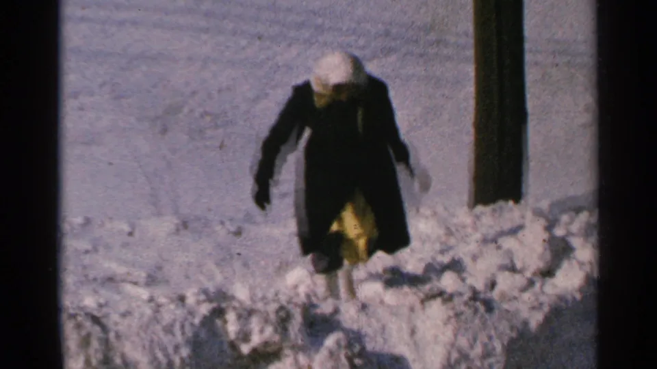 The woman continues through deep snow in 1965 Minnesota, her light head covering and yellow garment visible beneath a dark coat.