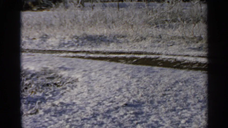 1965 Minnesota 8mm frame of snowy rural ground with brush and a dark curved pond edge or track crossing the scene.