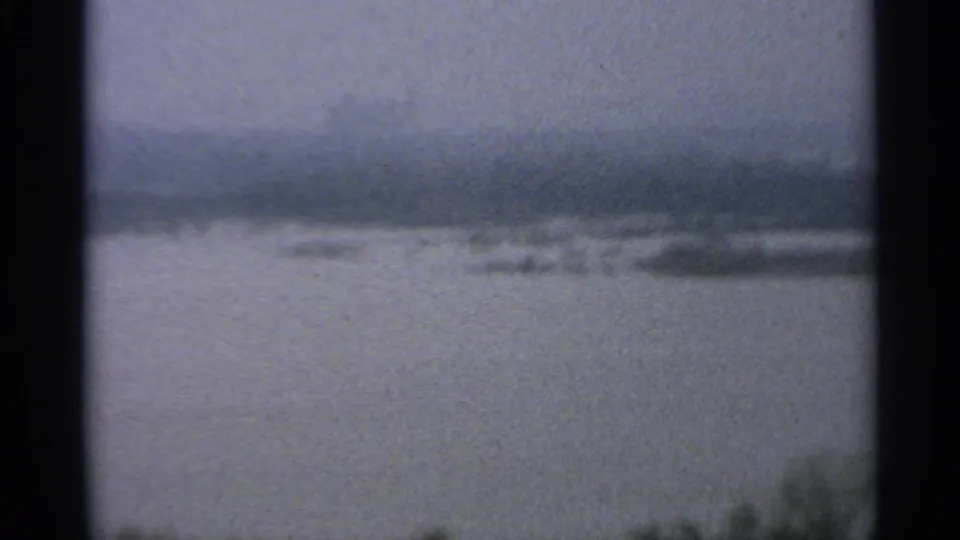 Muted 8mm view of a broad water surface under gray sky, with a low distant shoreline and dark foreground vegetation.
