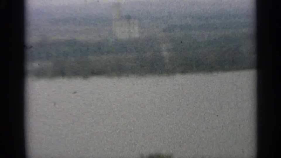A 1965 Minnesota 8mm frame showing pale water in the foreground and a blurred, wooded shoreline under overcast light.