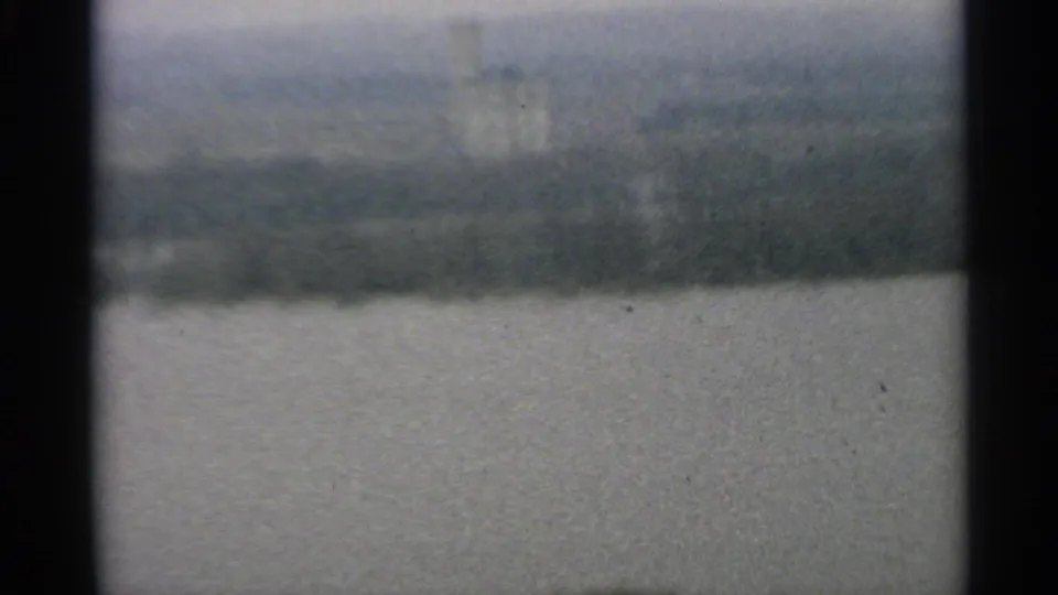1965 Minnesota home-movie frame with open water, a muted far shoreline, and hazy sky in grainy 8mm texture.