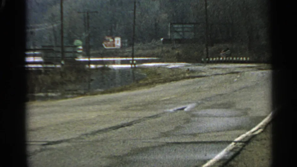 1965 Minnesota 8mm frame with a quiet road, pale shoulder, utility poles, and distant signs seen from a car.