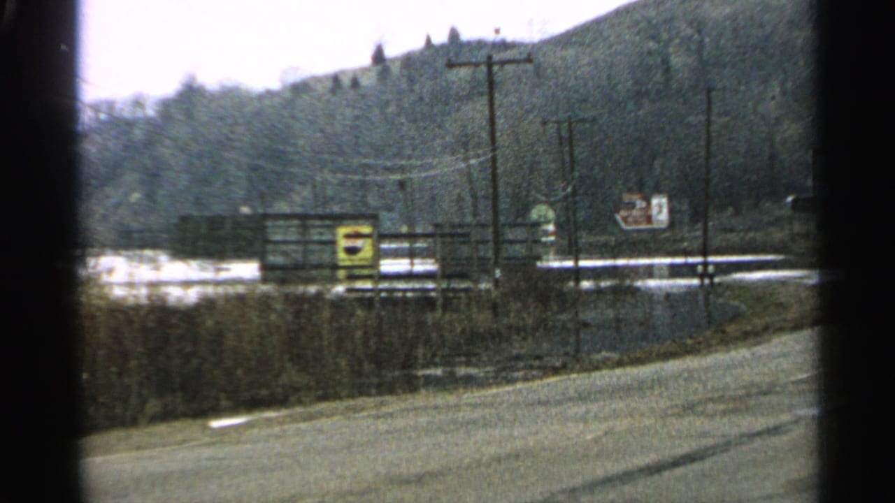 Rural roadside with utility poles, signs, water, and wooded hillside beyond.