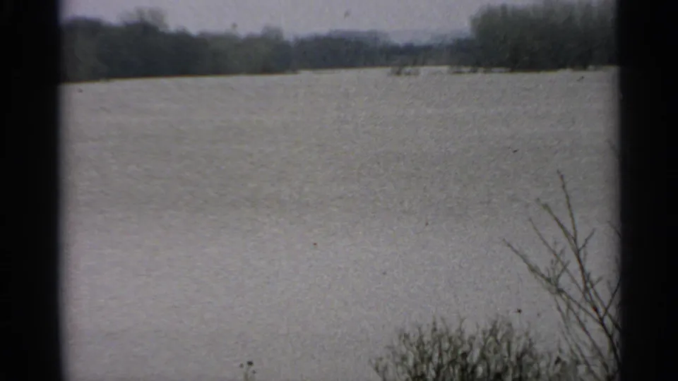1965 Minnesota 8mm frame of a gray lake under overcast light, with bare shrubs and a distant wooded shore.