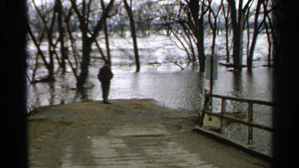 1965 Minnesota 8mm frame of a distant man standing by a flooded wooded area on an overcast morning.