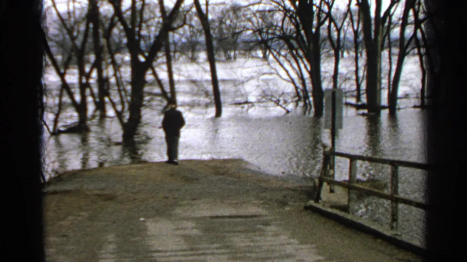 1965 Minnesota home-movie frame showing a man facing floodwater and leafless trees from a small path.