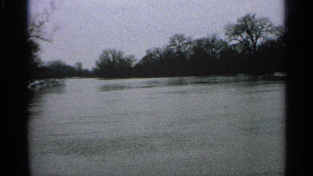 1965 Minnesota home-movie view across floodwater toward a dark treeline beneath a gray sky.