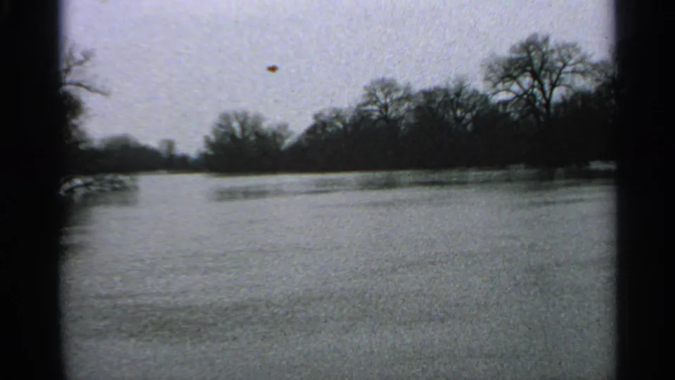 Soft 8mm frame from 1965 Minnesota showing murky floodwater, leafless trees, and a distant wooded bank.