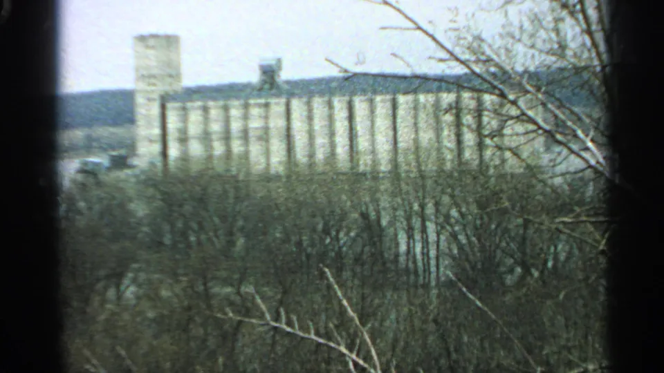 1965 home-movie frame with a grain-elevator-like building framed by leafless Minnesota woods.
