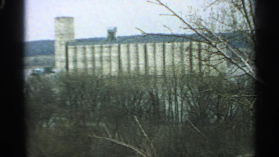 A wider 1965 Minnesota 8mm view of a distant elevator-like complex behind winter woods.