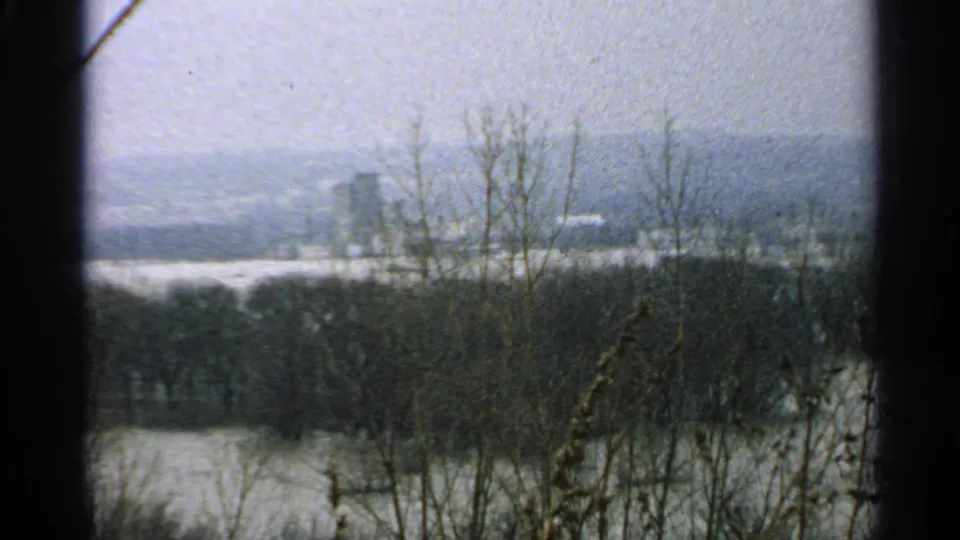 The 1965 Minnesota home-movie frame looks across water and winter-bare vegetation toward indistinct buildings on the far shore.