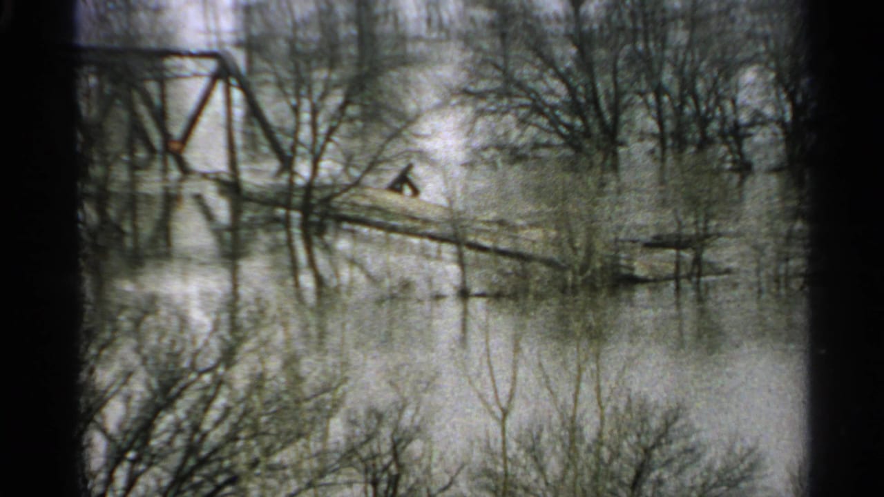 1965 Minnesota 8mm frame showing high water among bare trees, with a distant plank crossing and iron bridge structure beyond.