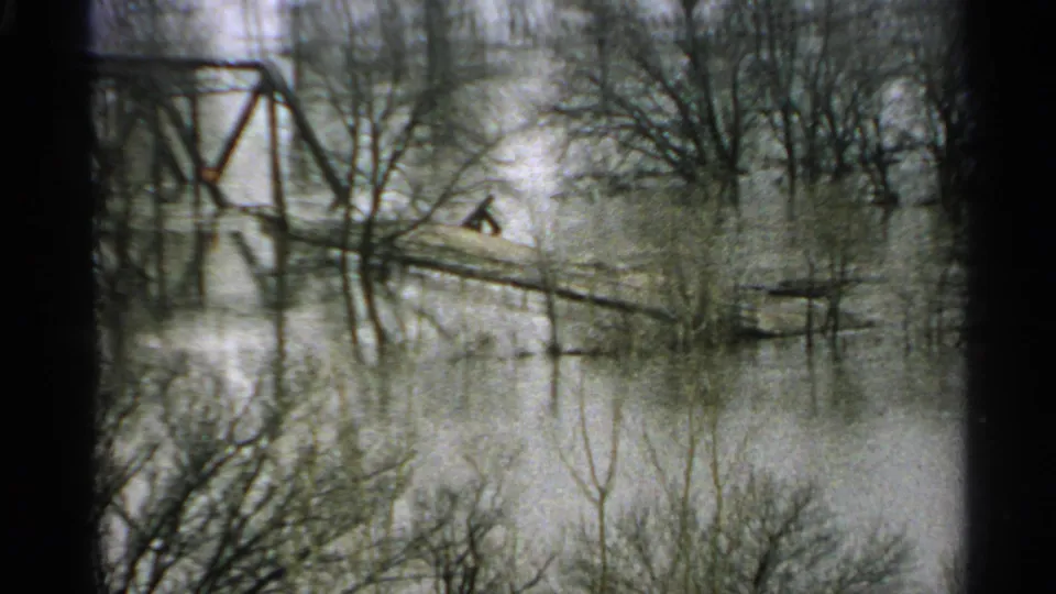 1965 Minnesota 8mm frame showing high water among bare trees, with a distant plank crossing and iron bridge structure beyond.