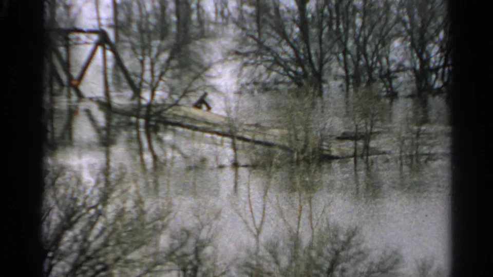 1965 Minnesota home-movie frame with a distant person crossing a narrow plank above floodwater near bare trees and an iron bridge.