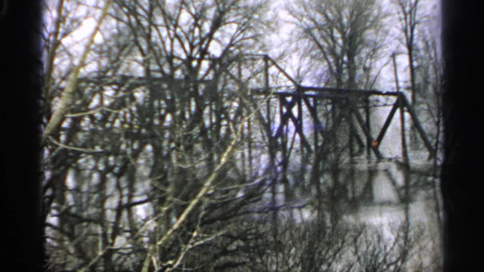 1965 Minnesota 8mm view of an iron truss bridge spanning floodwater, partly obscured by leafless foreground branches.