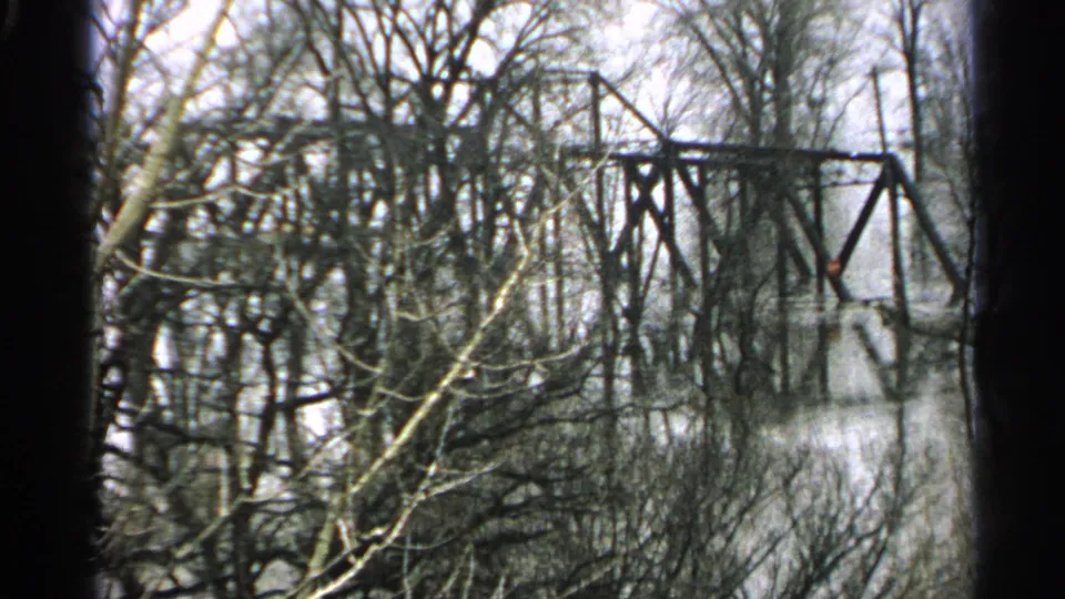 1965 Minnesota home-movie frame showing the iron truss bridge across a flooded river, framed by winter-bare trees.