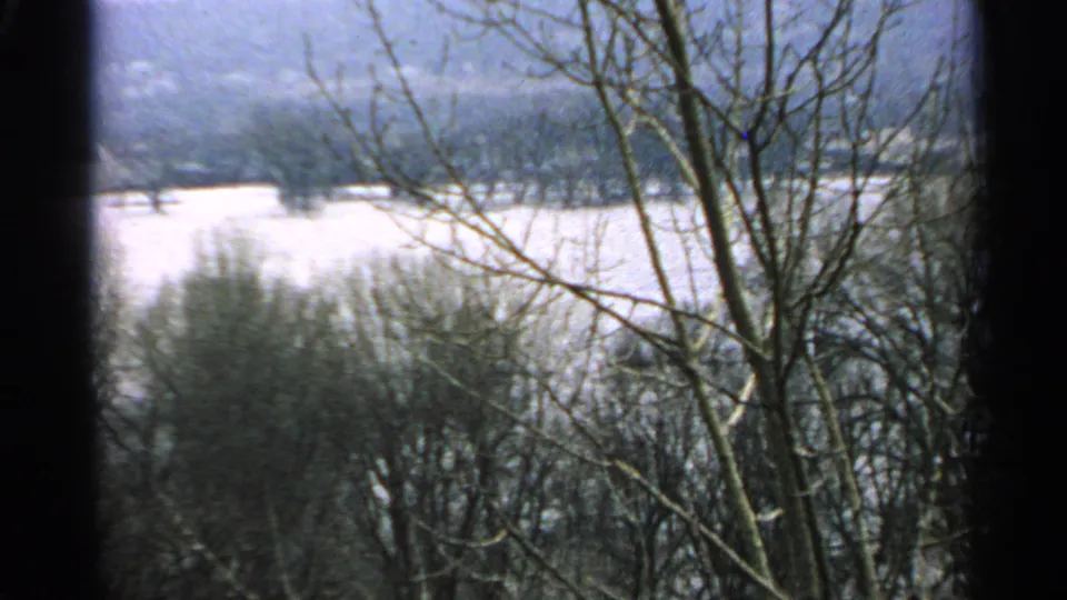 1965 Minnesota 8mm frame showing leafless trees in the foreground, a snowy open field, and wooded hills in the distance.