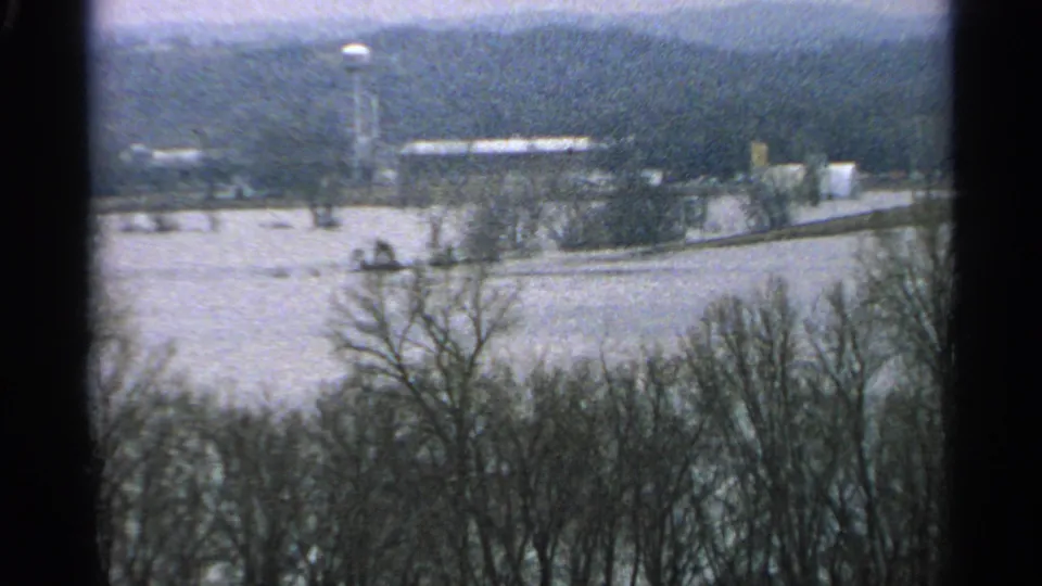 1965 Minnesota 8mm view across a snowy field toward bare trees, low buildings, a road or lane, and a water tower.
