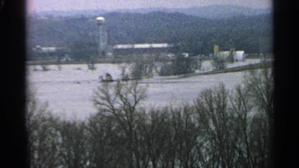 1965 Minnesota winter frame showing a water tower, low buildings, bare trees, and snow-covered ground from an elevated home-movie viewpoint.
