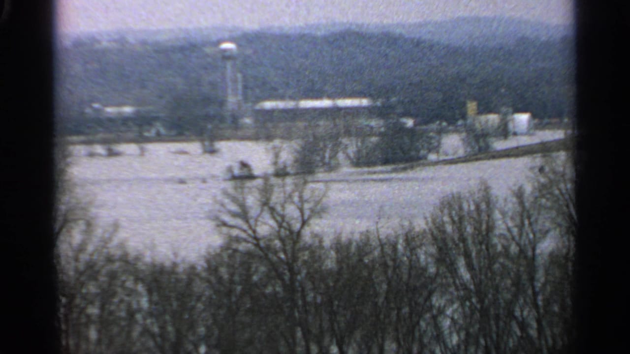 1965 Minnesota 8mm frame showing floodwater across low ground, bare trees in the foreground, distant buildings, and a water tower.
