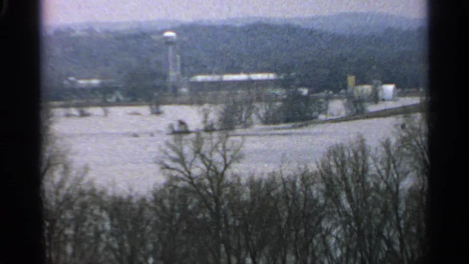 1965 Minnesota 8mm frame showing floodwater across low ground, bare trees in the foreground, distant buildings, and a water tower.