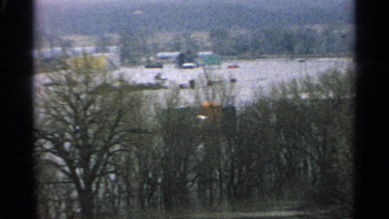 1965 Minnesota flood scene with leafless trees in front of a broad flooded area, low buildings, and scattered dark objects in the water.