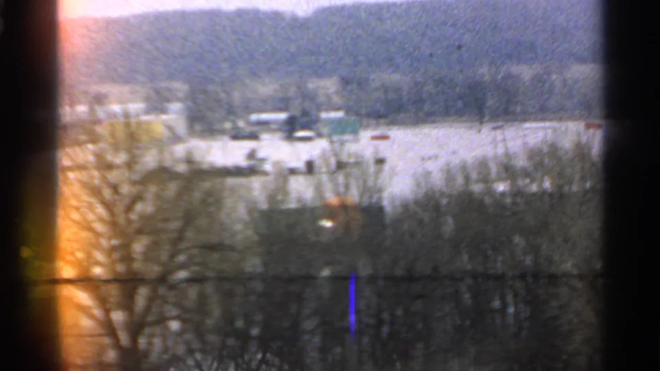1965 Minnesota 8mm frame of a flooded rural area viewed past bare trees, with low structures and a warm film light leak at the edge.