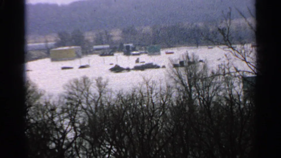 1965 Minnesota home-movie frame showing a wide flooded landscape, dark foreground trees, and small structures or debris across the water.